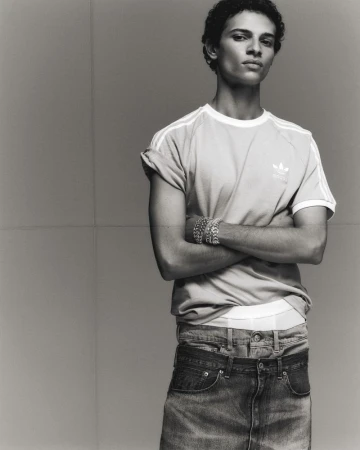 Black-and-white portrait of an adult male with curly hair, wearing an Adidas T-shirt and jeans, posing with arms crossed indoors.