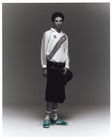 Full-length portrait of a male model wearing an Adidas white sports jersey, black shorts, and green sneakers against a plain grey studio background.
