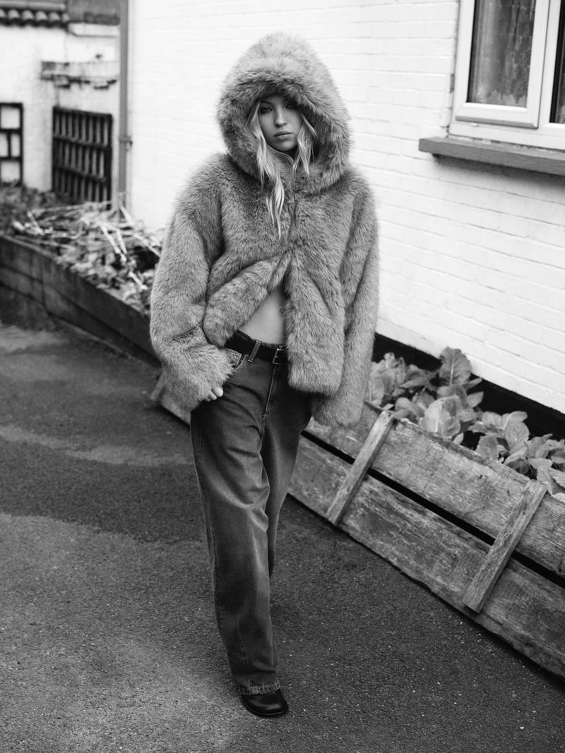 Black and white fashion shot of a blonde adult female wearing a fur coat and jeans walking on an outdoor city street.