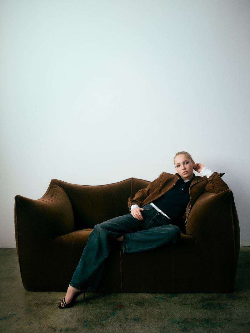 A blond woman in a brown jacket and jeans reclines on a brown couch in a relaxed pose against a white wall backdrop indoors.