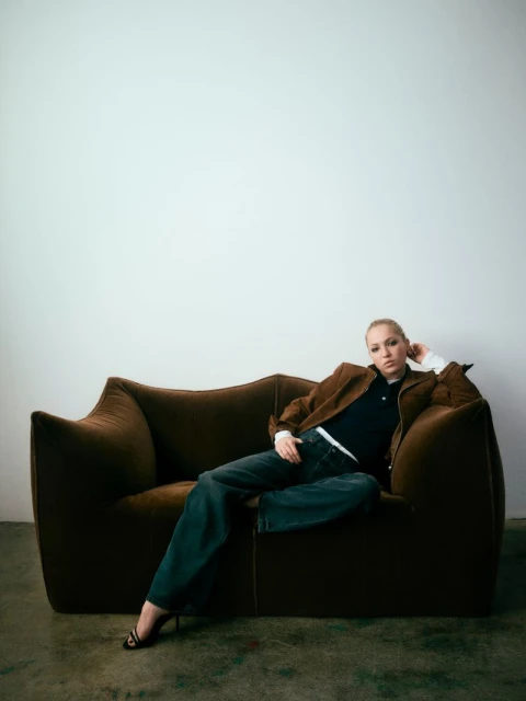 A blond woman in a brown jacket and jeans reclines on a brown couch in a relaxed pose against a white wall backdrop indoors.