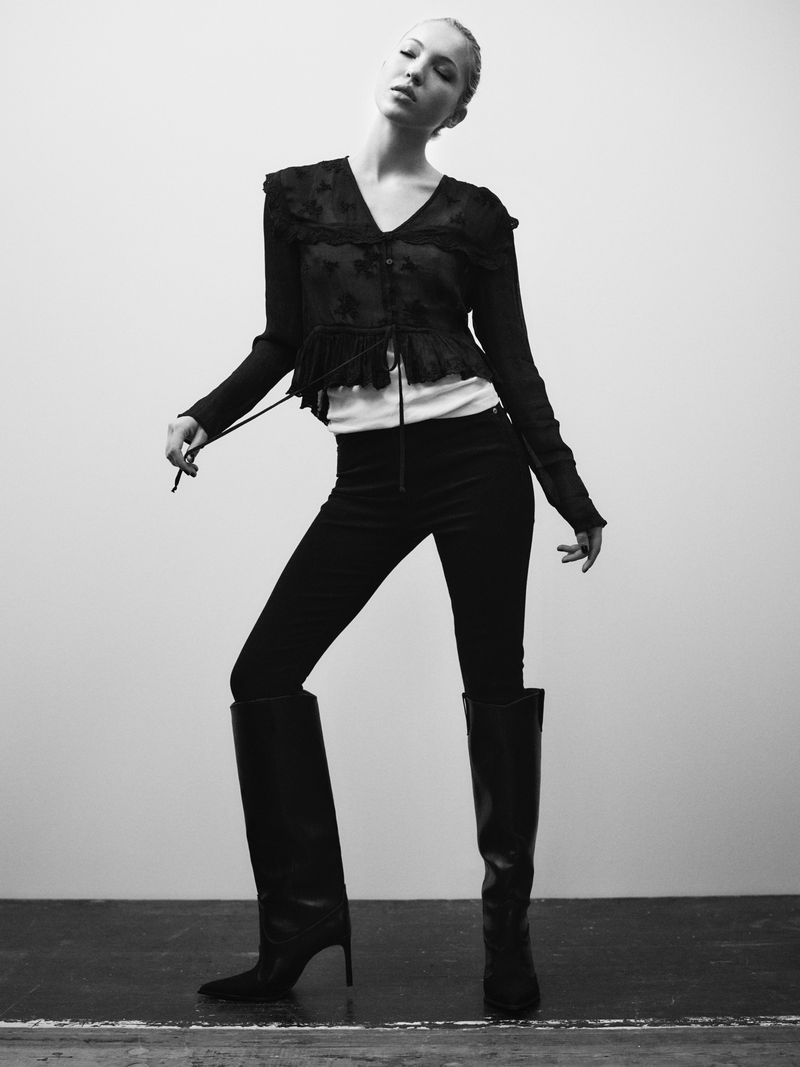 Full-length black-and-white portrait of a posing adult woman in a lace top, dark jeans, and tall boots against a neutral backdrop.