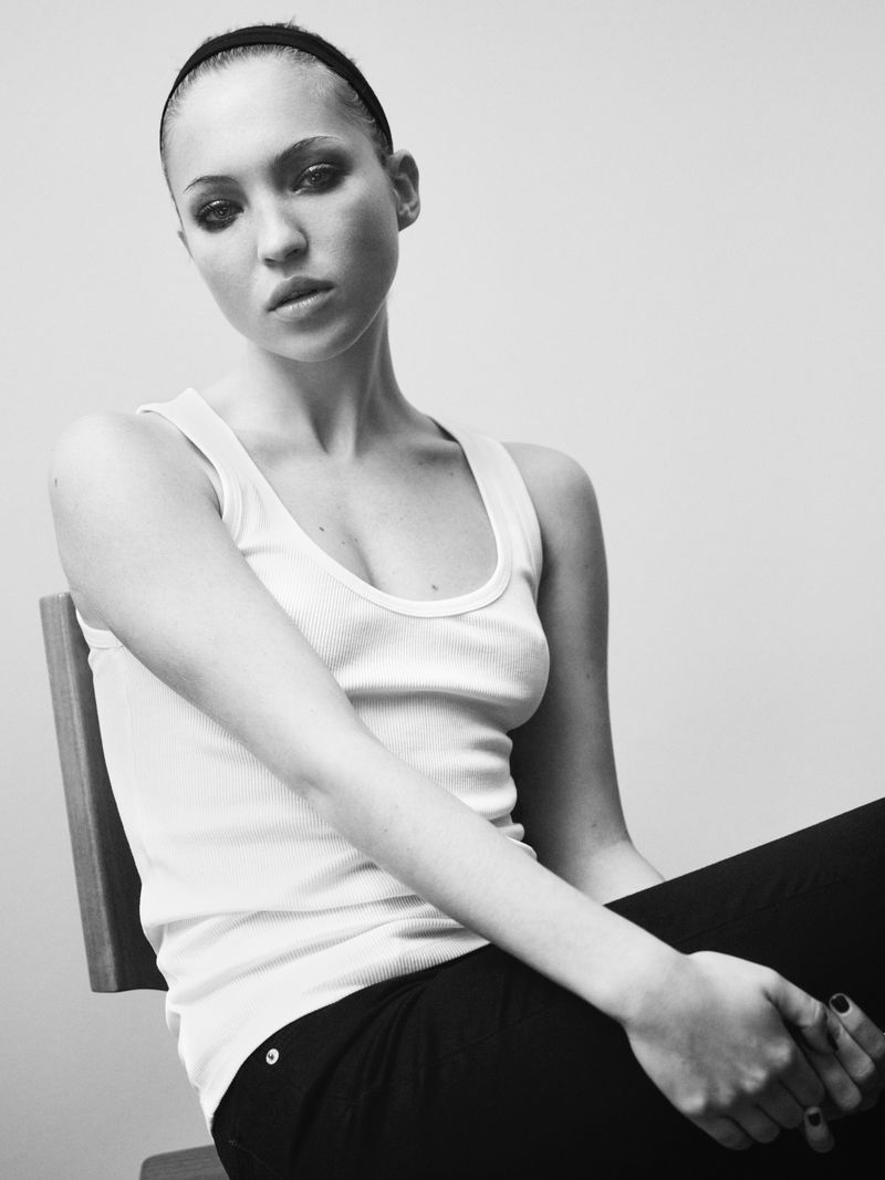Black and white portrait of a woman with dark hair wearing a tank top and dark pants, sitting on a chair in a bright studio.