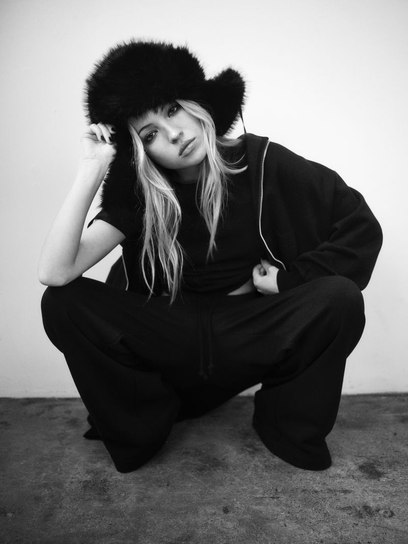 A young woman with long blonde hair squats indoors, wearing a black outfit and furry hat, set against a neutral wall in a portrait.