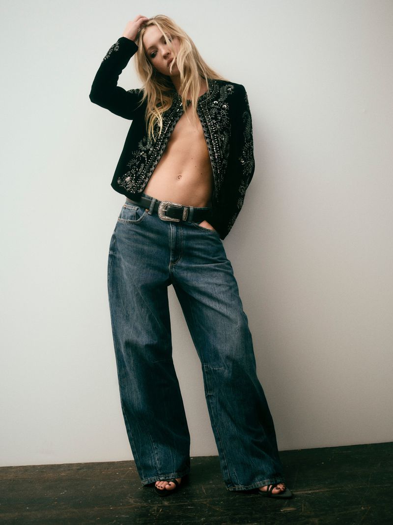 Model Lila Moss wearing wide-leg denim jeans and an embroidered black jacket against a plain white studio background.