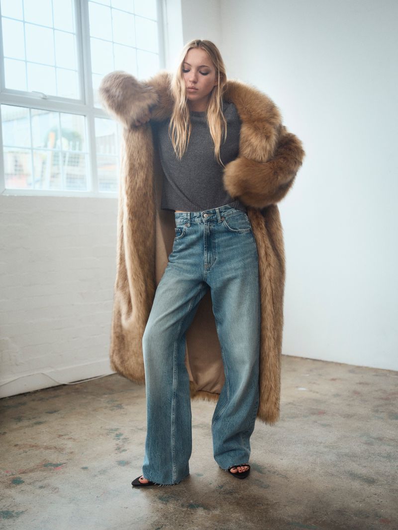 Woman poses in long faux fur coat, gray top, and blue jeans in a minimalist setting, bathed in soft natural light from nearby windows.