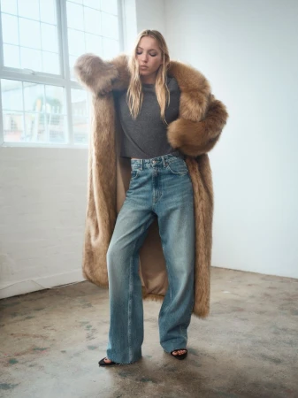 Woman poses in long faux fur coat, gray top, and blue jeans in a minimalist setting, bathed in soft natural light from nearby windows.
