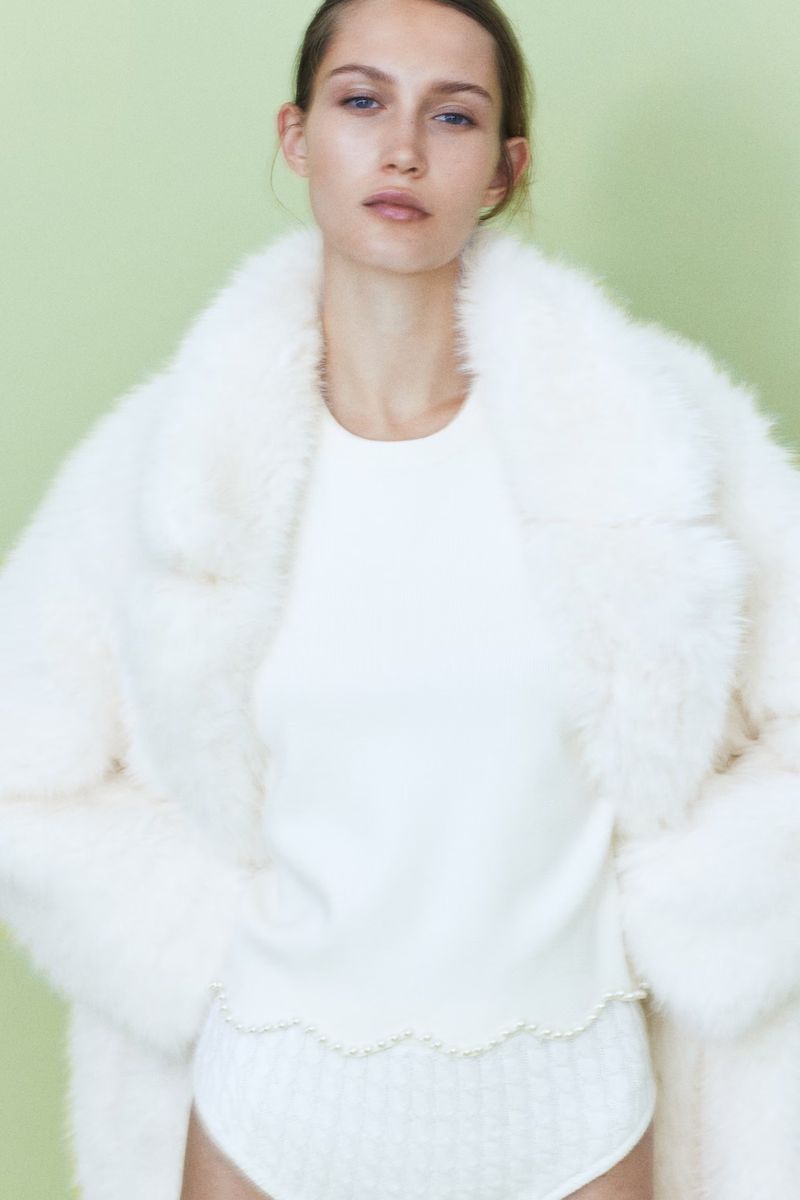 An adult female model poses indoors wearing a white sweater, briefs, and a faux fur coat against a light green backdrop.