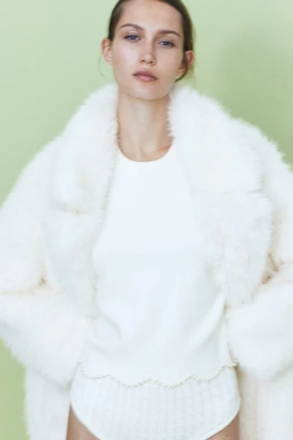 An adult female model poses indoors wearing a white sweater, briefs, and a faux fur coat against a light green backdrop.