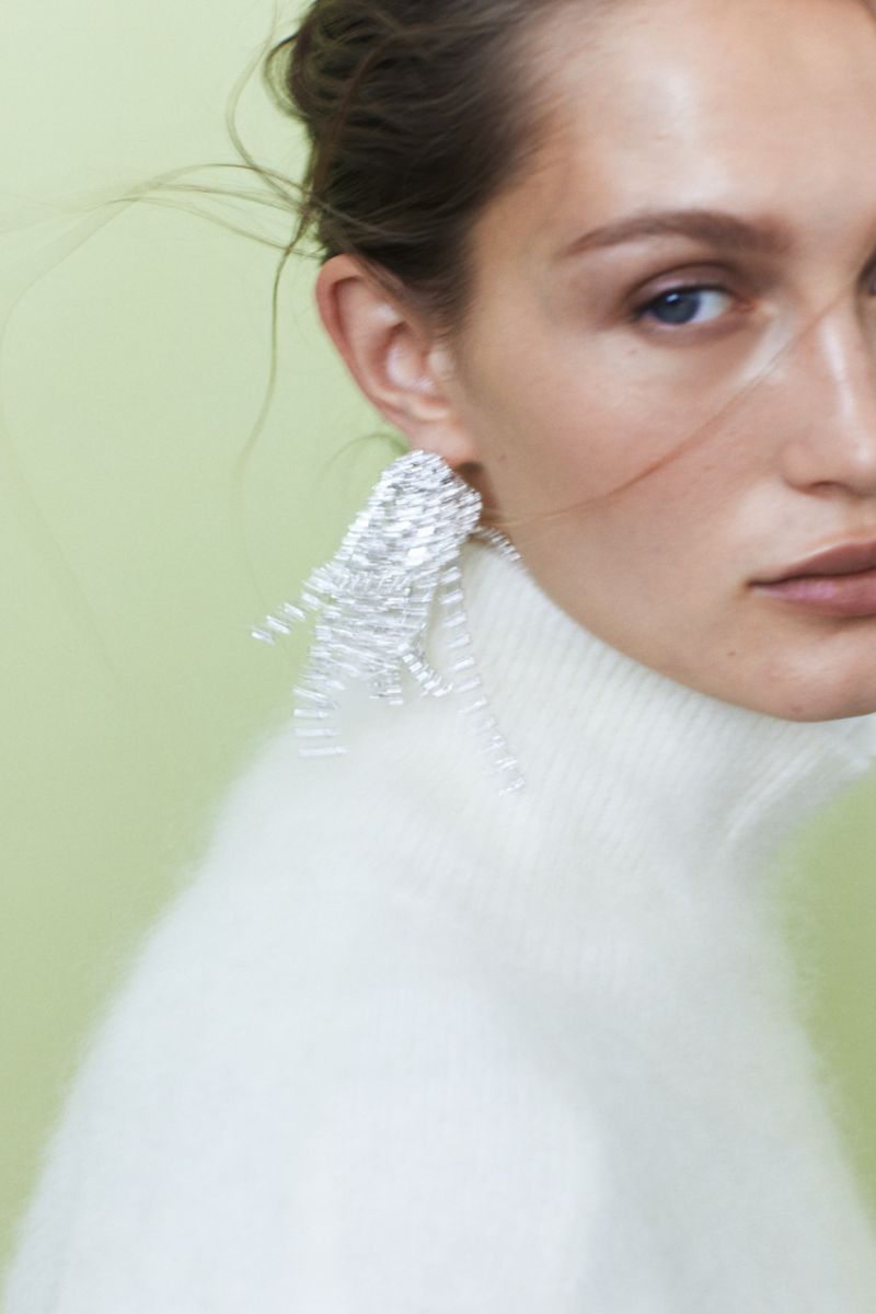 Close-up of a woman with fair skin, light eyes, and silver geometric earrings, wearing a white turtleneck sweater, set against a light green background.