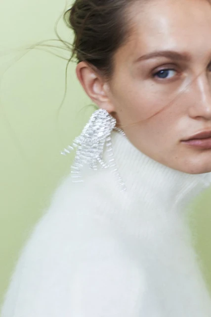 Close-up of a woman with fair skin, light eyes, and silver geometric earrings, wearing a white turtleneck sweater, set against a light green background.