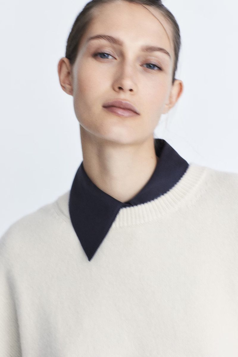 Portrait of a female model wearing a cream knit sweater over a dark collared shirt against a white backdrop.