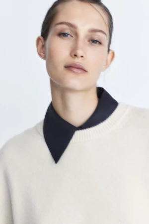 Portrait of a female model wearing a cream knit sweater over a dark collared shirt against a white backdrop.