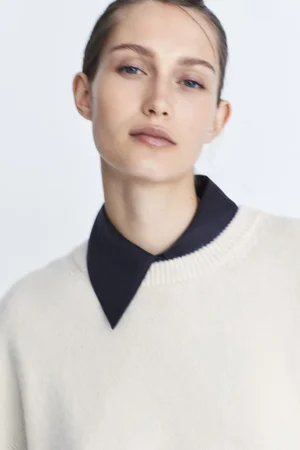 Portrait of a female model wearing a cream knit sweater over a dark collared shirt against a white backdrop.