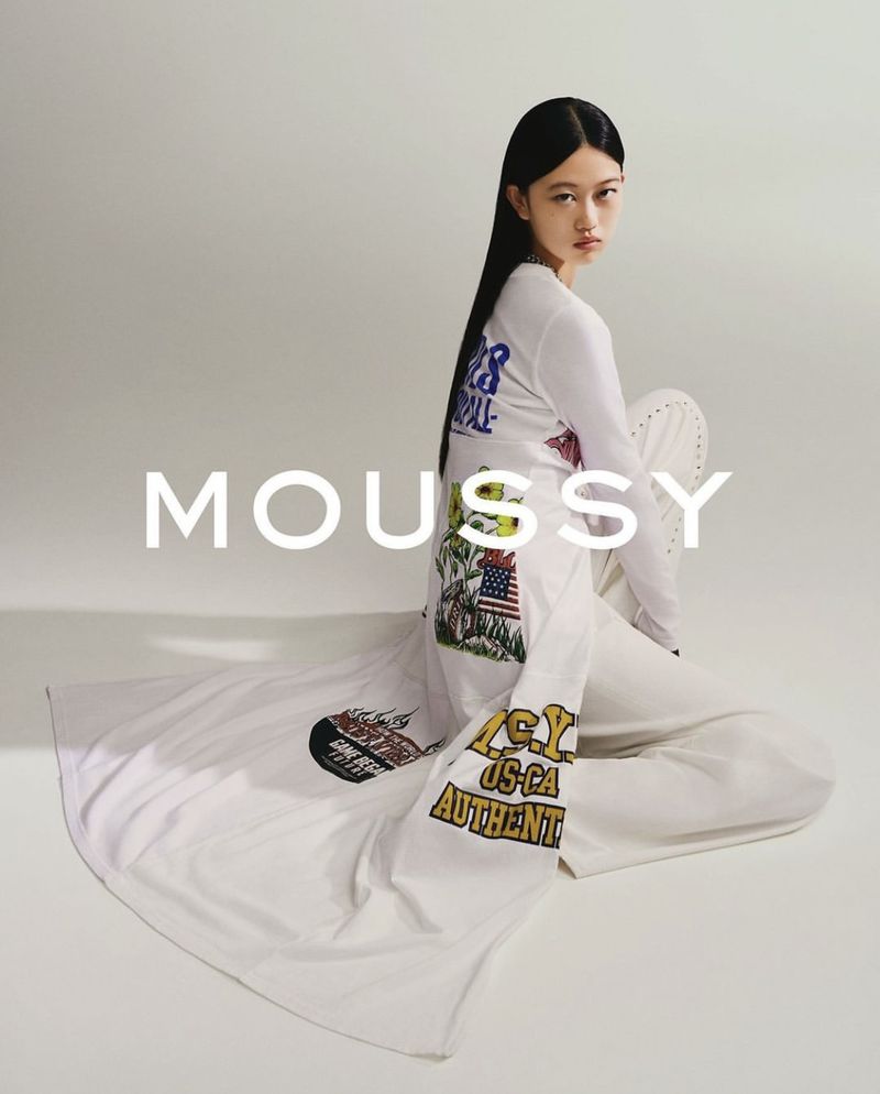 Model with long dark hair wearing a white printed long-sleeve top and white trousers sitting against a grey background with MOUSSY text overlay
