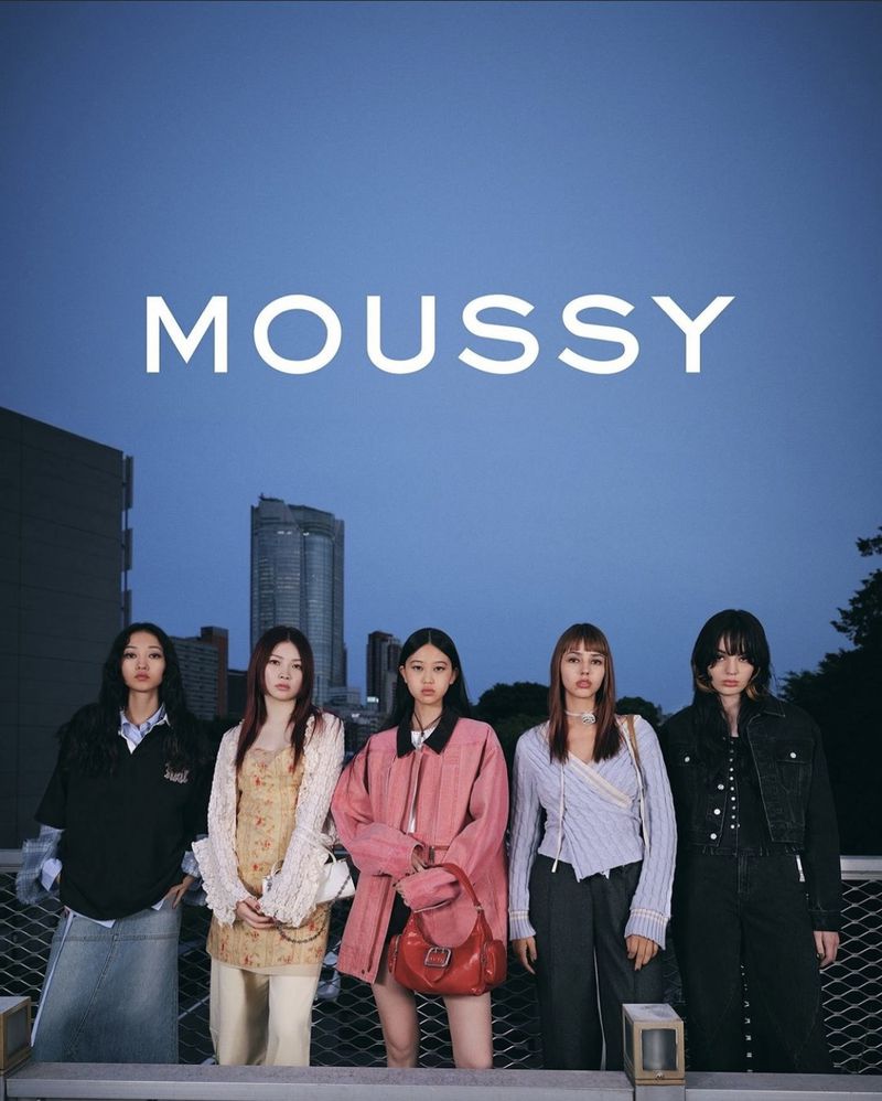 Five young Asian women pose outdoors against a city backdrop in Moussy apparel. Fashion-forward clothing in muted tones dominates.