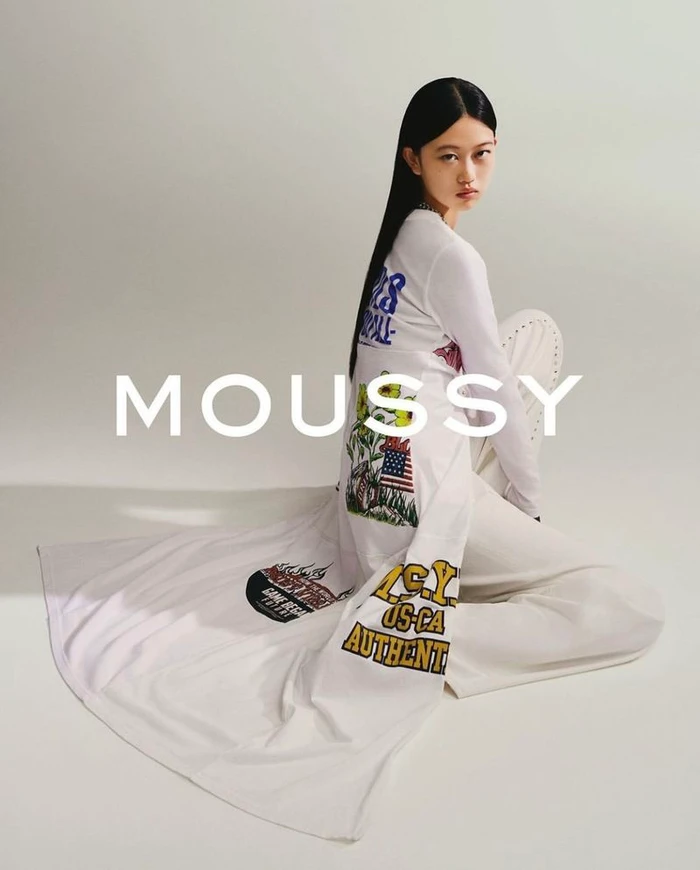 Model with long dark hair wearing a white printed long-sleeve top and white trousers sitting against a grey background with MOUSSY text overlay