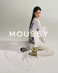 Model with long dark hair wearing a white printed long-sleeve top and white trousers sitting against a grey background with MOUSSY text overlay