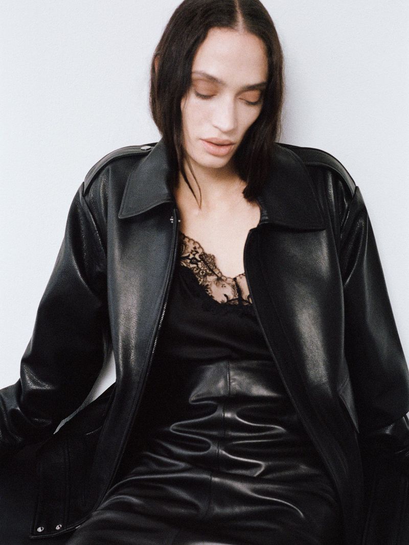 A woman with long dark hair poses in a studio wearing a black leather trench coat and pants, with a black lace top underneath.