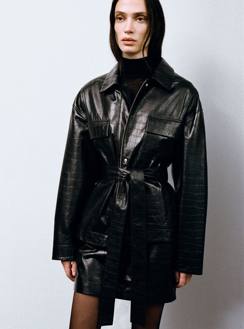 Indoor shot of a slender adult with black hair wearing a black turtleneck and a belted black faux-crocodile jacket dress over tights.