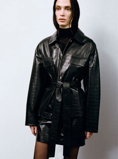 Indoor shot of a slender adult with black hair wearing a black turtleneck and a belted black faux-crocodile jacket dress over tights.