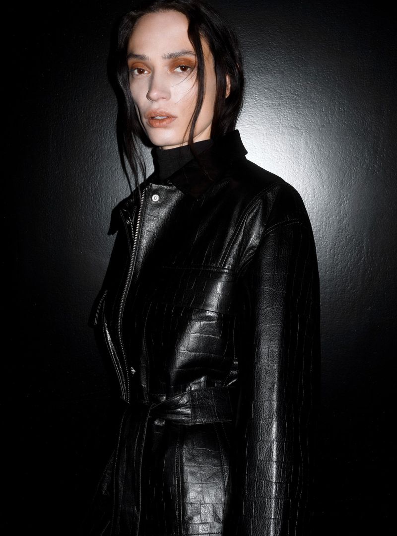 Androgynous model with long dark hair posing in a black faux crocodile leather coat against a dark grey backdrop in a studio shot.