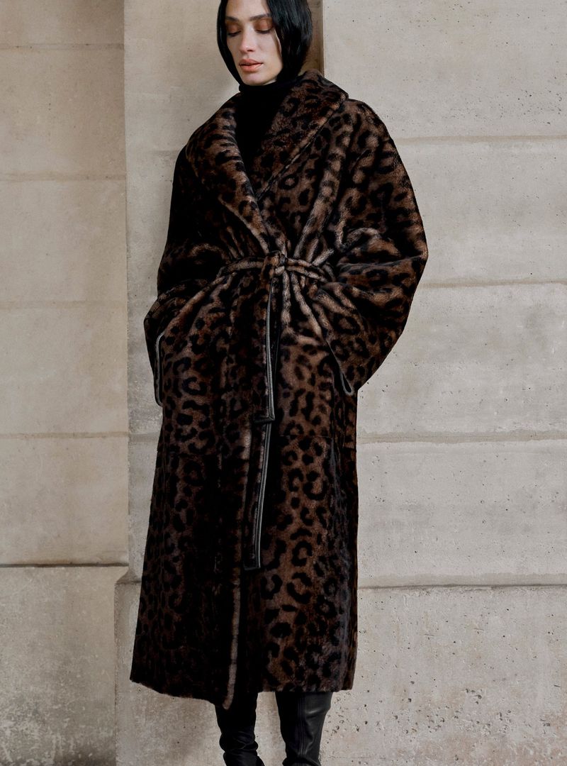Model wearing a long belted leopard print faux fur coat and black boots standing against a stone building exterior.