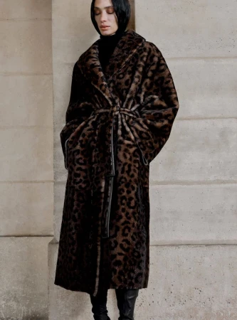 Model wearing a long belted leopard print faux fur coat and black boots standing against a stone building exterior.