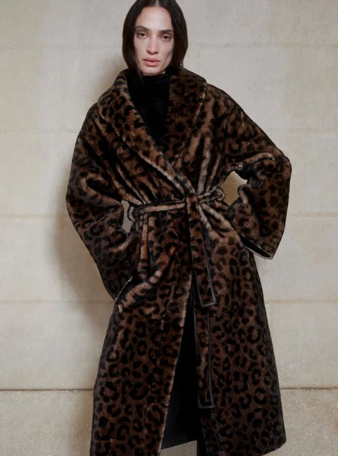 Elegant adult woman in a long, spotted faux fur coat with a belt, posing indoors against a neutral wall. Fashion shot, full view.