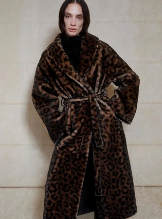Elegant adult woman in a long, spotted faux fur coat with a belt, posing indoors against a neutral wall. Fashion shot, full view.
