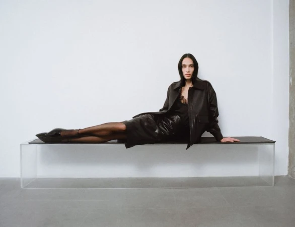 Model wearing a dark leather jacket and skirt reclining on a clear glass bench against a plain white wall