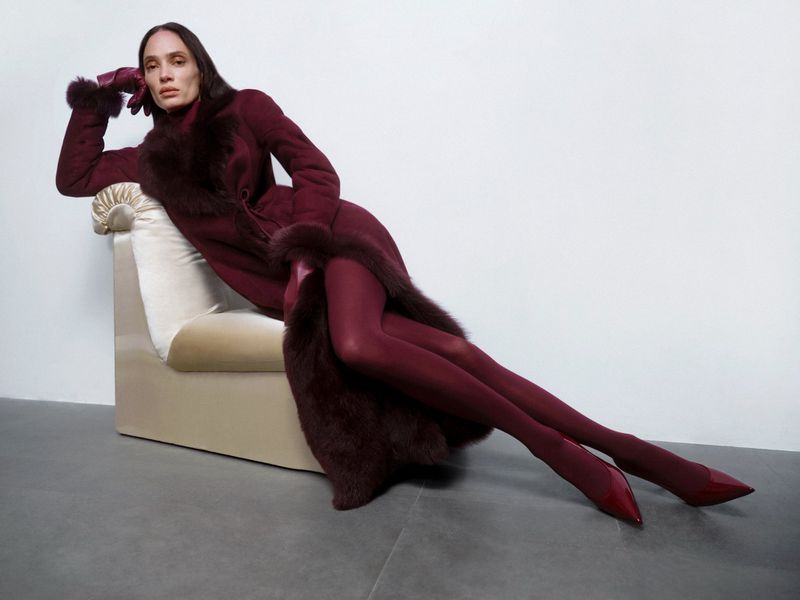 A woman in a fur-trimmed coat poses on a chair in a fashion studio. She wears matching tights and shoes, styled with gloves.