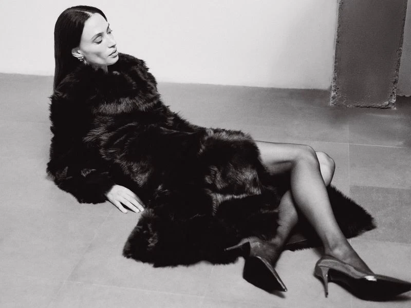 Elegant woman in a fur coat poses on the floor in black heels and black tights against a white background, showcasing fashion.