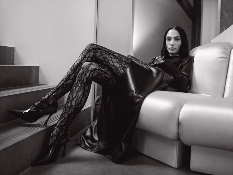 A woman in a long, black, shiny coat and lace tights poses on a white couch near steps in an indoor, monochrome fashion shot.