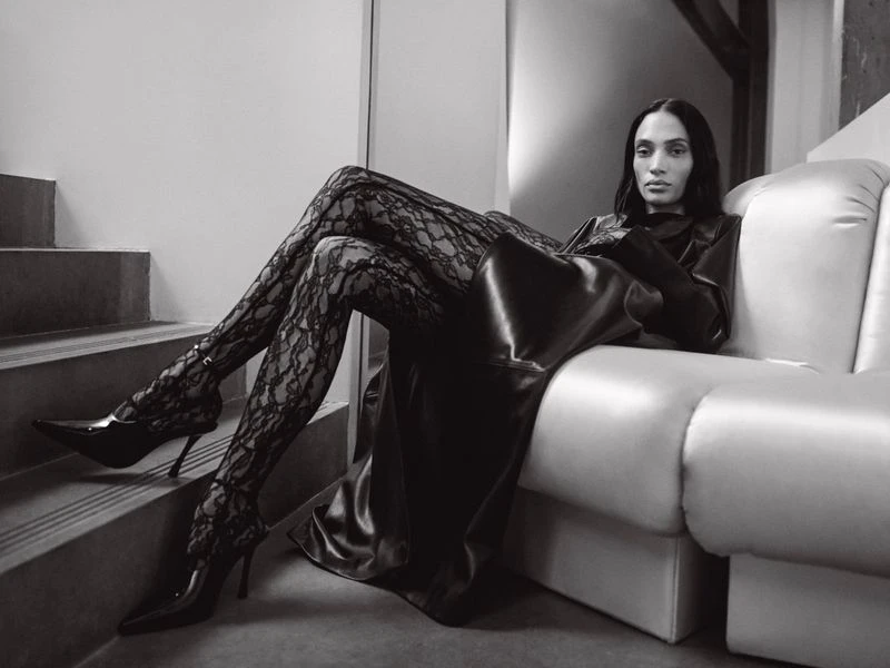 A woman in a long, black, shiny coat and lace tights poses on a white couch near steps in an indoor, monochrome fashion shot.