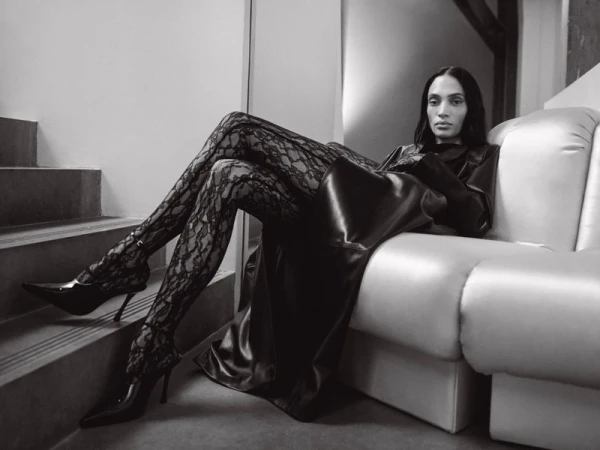 A woman in a long, black, shiny coat and lace tights poses on a white couch near steps in an indoor, monochrome fashion shot.
