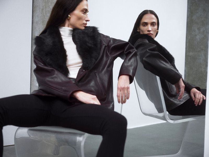 Brunette woman in black leather jacket with fur collar, sitting on white chair in a studio setting with neutral backdrop, fashion editorial.