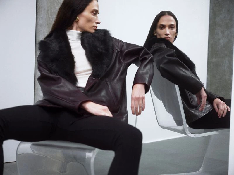Brunette woman in black leather jacket with fur collar, sitting on white chair in a studio setting with neutral backdrop, fashion editorial.