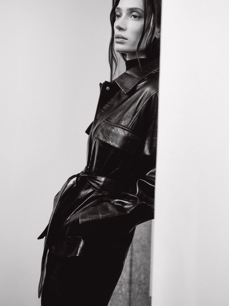 Monochrome fashion portrait of a model in a dark belted leather jacket leaning against a pale wall.