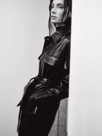 Monochrome fashion portrait of a model in a dark belted leather jacket leaning against a pale wall.