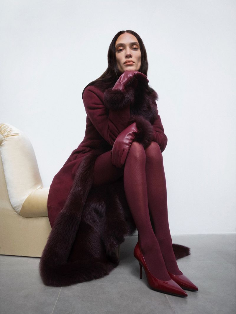 Elegant woman in a burgundy shearling coat with fur trim, burgundy leggings, and matching heels poses against a white background.