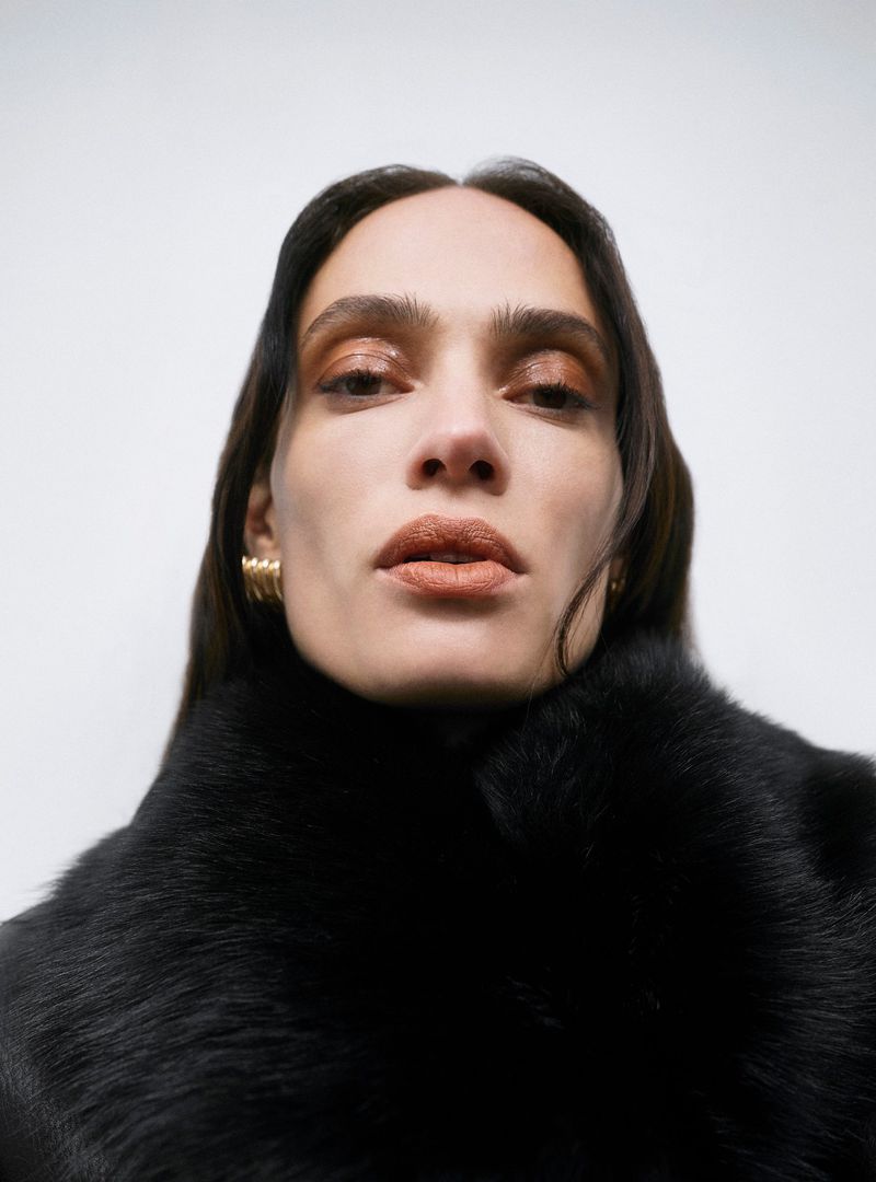 Close-up portrait of an adult female with long brown hair wearing a black fur collar and gold earrings against a white background.