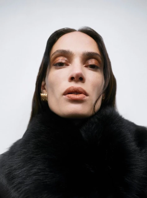 Close-up portrait of an adult female with long brown hair wearing a black fur collar and gold earrings against a white background.