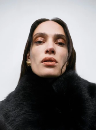 Close-up portrait of an adult female with long brown hair wearing a black fur collar and gold earrings against a white background.
