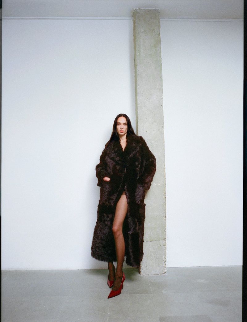 Model wearing a dark brown furry coat and red heels posing against a white wall next to a concrete pillar.