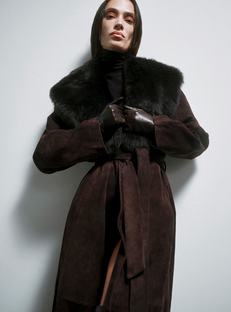 A brown-haired woman models a brown suede coat with fur collar against a light wall. She wears black gloves and a black turtleneck.