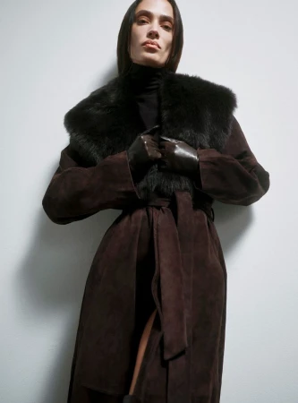 A brown-haired woman models a brown suede coat with fur collar against a light wall. She wears black gloves and a black turtleneck.