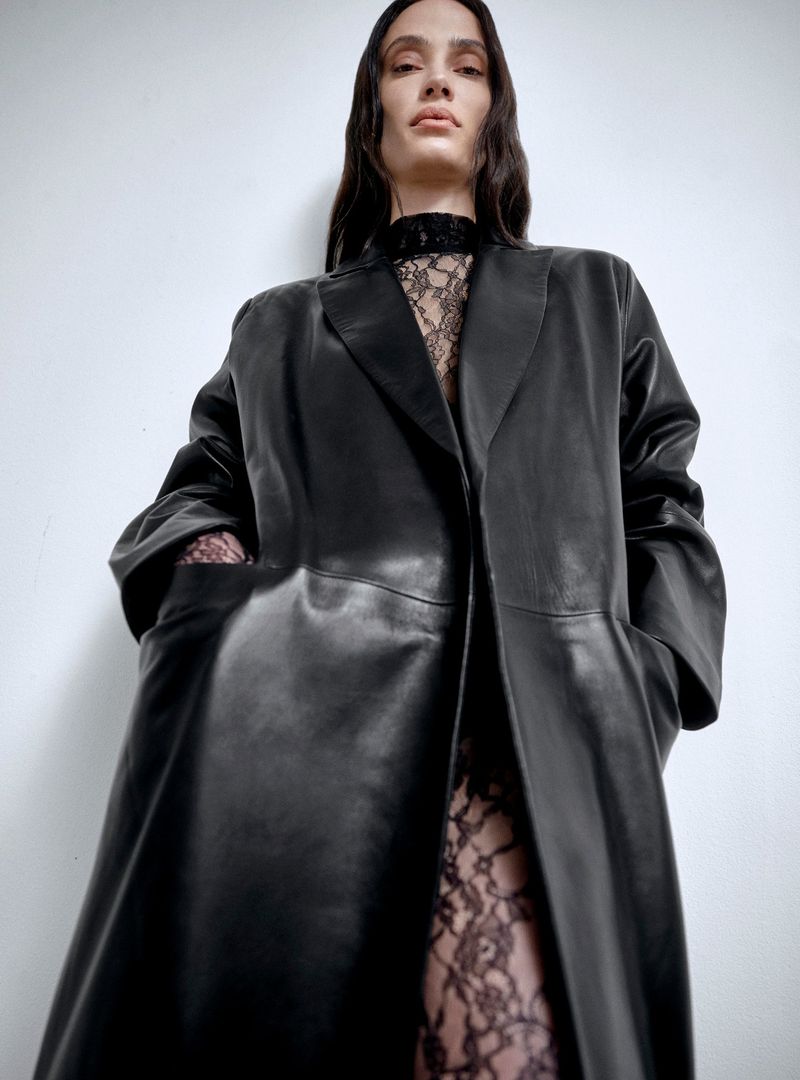 A fashionable adult woman with long hair wears a long black leather coat over a lace top and leggings against a plain white backdrop.