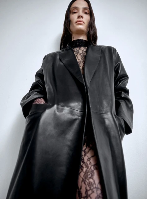 A fashionable adult woman with long hair wears a long black leather coat over a lace top and leggings against a plain white backdrop.