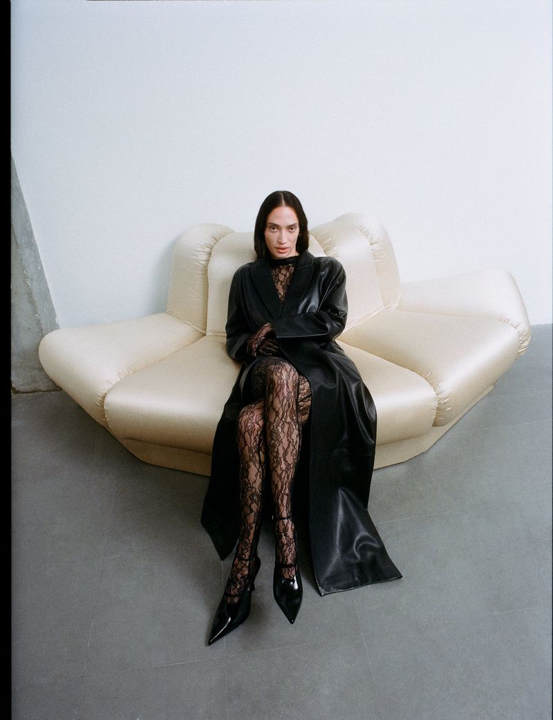 A woman in a black coat and floral tights sits on a cream-colored couch in front of a white wall, creating a fashion-forward indoor portrait.